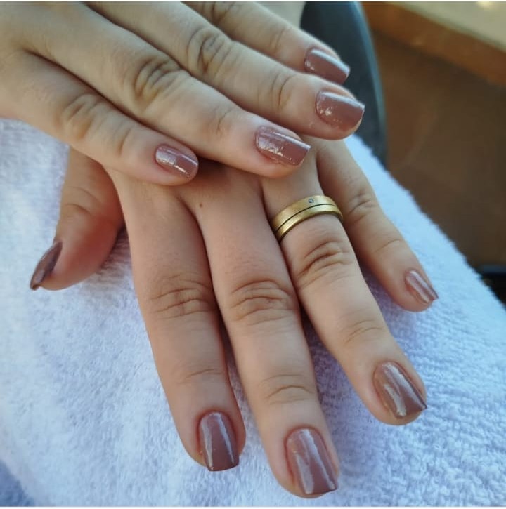 Mãos femininas com unhas naturais pintadas em tom nude brilhante, com um anel dourado na mão esquerda.
