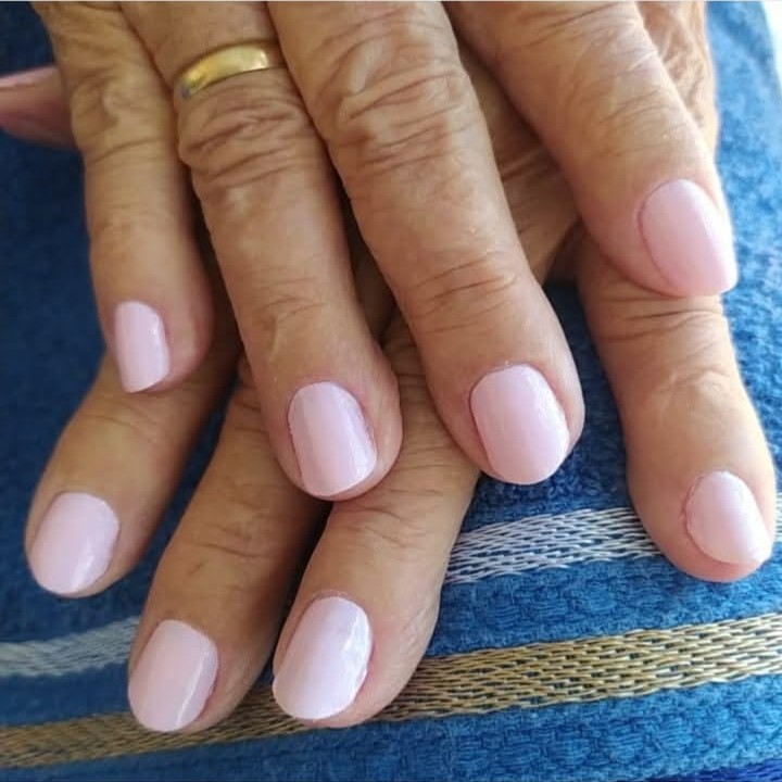 Mãos de uma pessoa idosa com unhas naturais pintadas de rosa claro, repousando sobre um tecido azul.